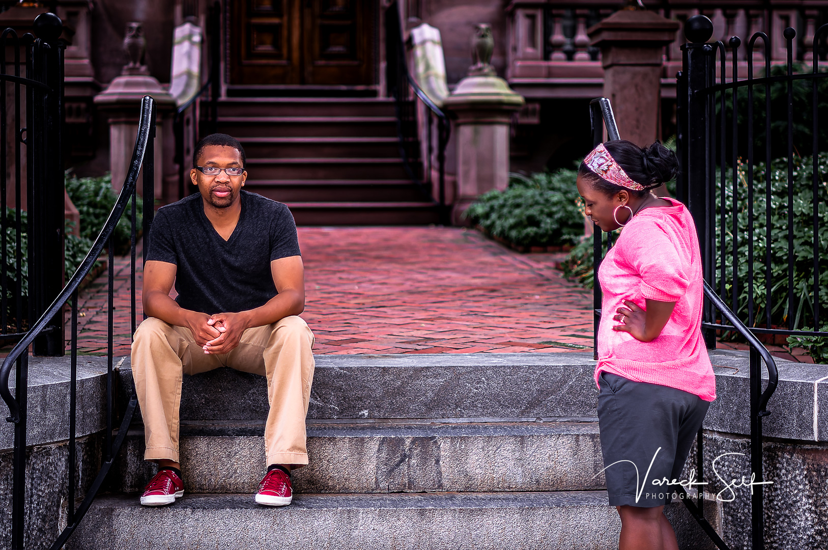 A couple shares an unscripted moment on stone steps outside a historic brick building — the man seated and looking across at the woman in pink, who stands with hand on hip, in quiet conversation between poses.