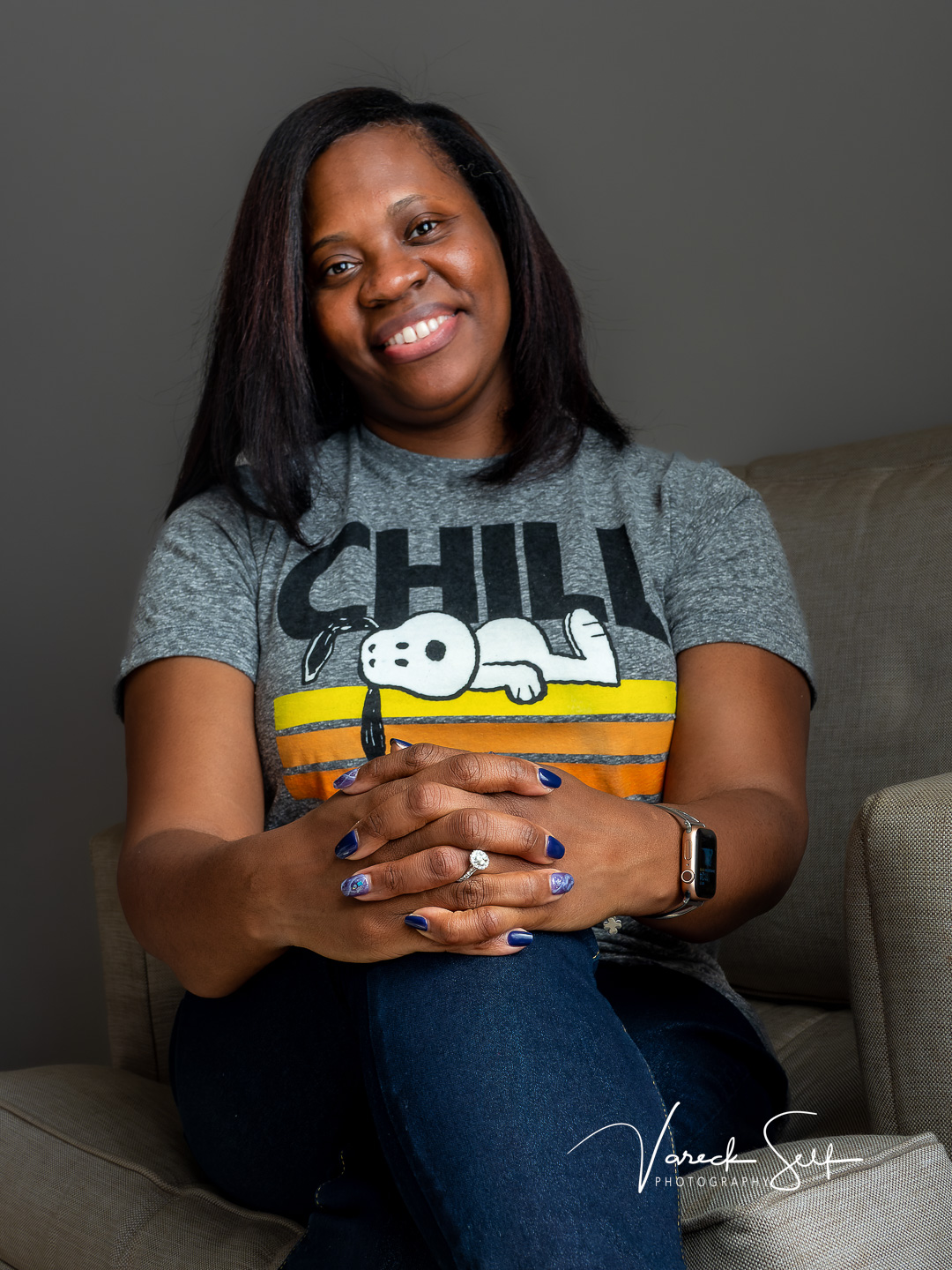 A woman smiles warmly at the camera, hands clasped in her lap displaying an engagement ring, wearing a Snoopy graphic tee, seated on a neutral couch against a soft gray background.
